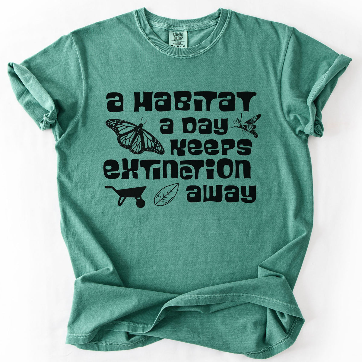 Light green "A Habitat a Day Keeps Extinction Away" t-shirt on Comfort Colors tee, featuring a monarch butterfly, clearwing moth, wheelbarrow, and leaf icon with bold black lettering. Gardener and conservation gift shirt for native plant and pollinator enthusiasts.