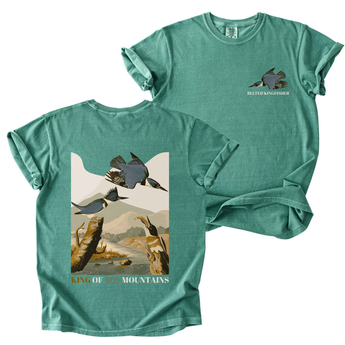 Light green "Belted Kingfisher, King of the Mountains" t-shirt on Comfort Colors tee, featuring a small front chest logo and a full illustrated back print of kingfishers over a river landscape in retro poster style. Birdwatcher gift shirt for wildlife and nature enthusiasts.