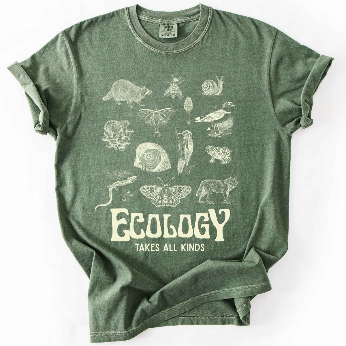 Moss "Ecology Takes All Kinds" t-shirt on Comfort Colors tee, featuring a grid of raccoons, owls, wolves, salamanders, mushrooms, moths, and other wildlife in cream ink. Nature lover and biology gift shirt for ecologists and wildlife enthusiasts.