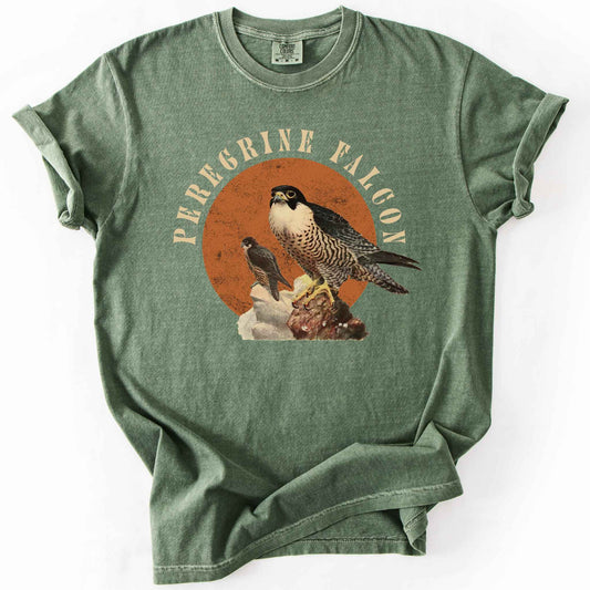Moss "Peregrine Falcon" t-shirt on Comfort Colors tee, featuring vintage style falcon illustration perched on a rock against an orange sun backdrop, with arched cream text. Birdwatcher and raptor lover gift shirt for falconry and wildlife fans.