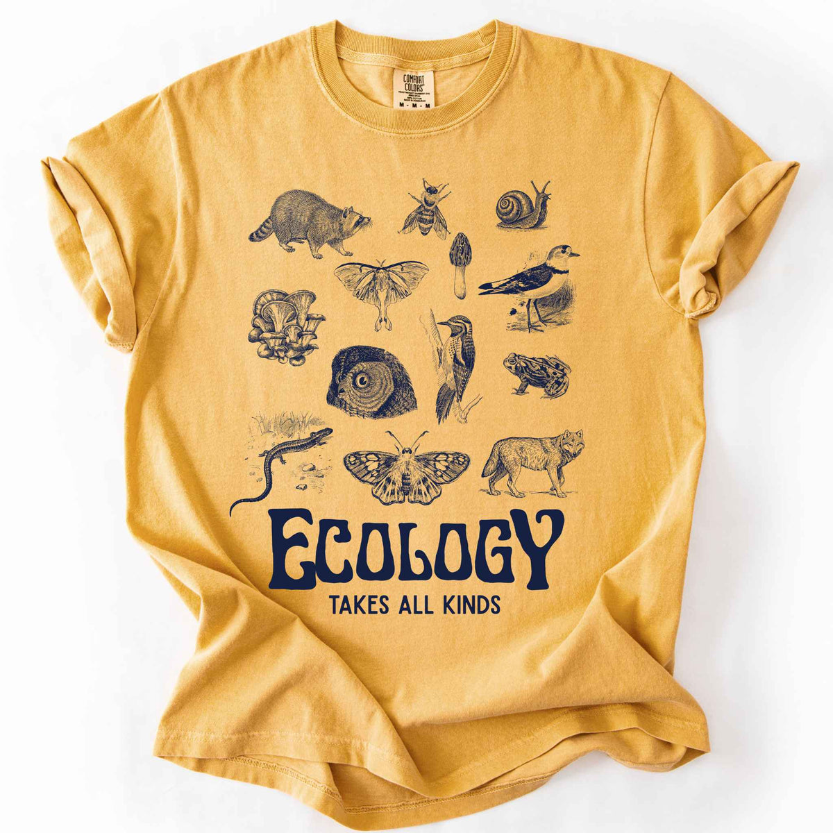 Mustard "Ecology Takes All Kinds" t-shirt on Comfort Colors tee, featuring a grid of raccoons, owls, wolves, salamanders, mushrooms, moths, and other wildlife in cream ink. Nature lover and biology gift shirt for ecologists and wildlife enthusiasts.
