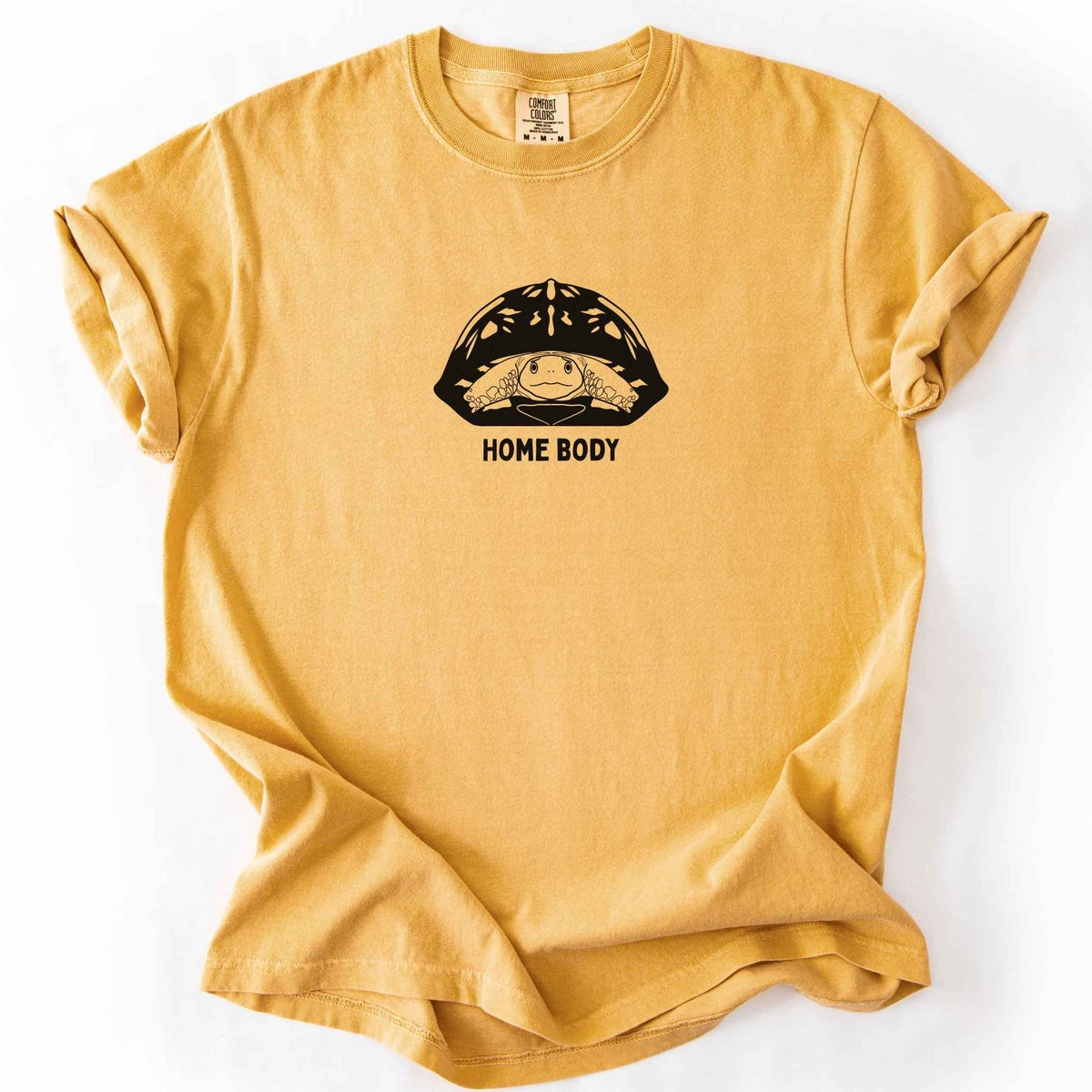 Mustard "Home Body" t-shirt on Comfort Colors tee, featuring a bold black graphic of a turtle peeking out from its shell above simple lettering. Funny turtle lover gift shirt for introverts and reptile enthusiasts.