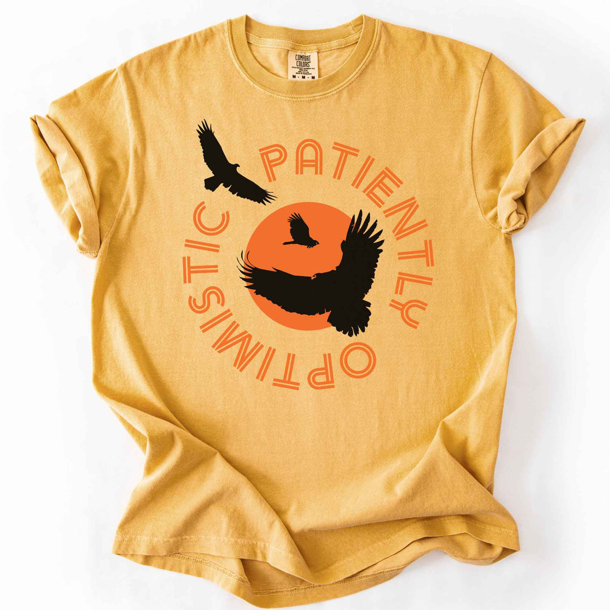 Mustard "Patiently optimistic turkey vulture" t-shirt on comfort colors tee. Silhouette turkey vulture graphics against an orange sun surrounded by curved text. Bird lover and wildlife enthusiast gift. 