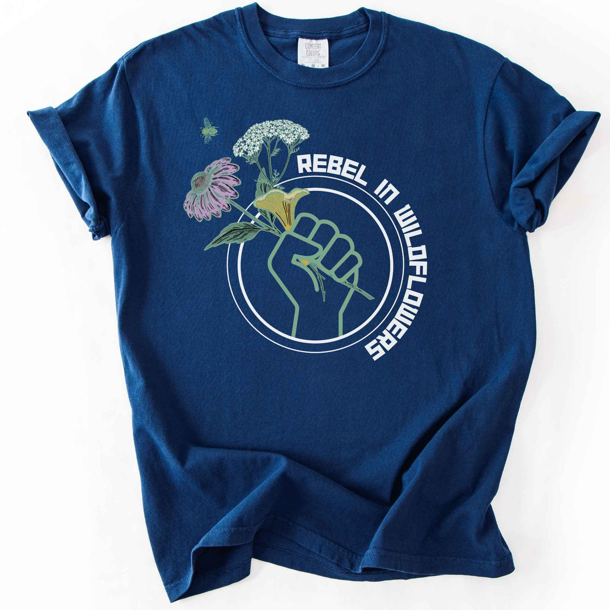 Navy "Rebel in Wildflowers" t-shirt on Comfort Colors tee, featuring a raised fist holding coneflower, yarrow, and a mushroom with a bumblebee in a circular badge design. Gardener and native plant gift shirt for wildflower and pollinator conservation fans.