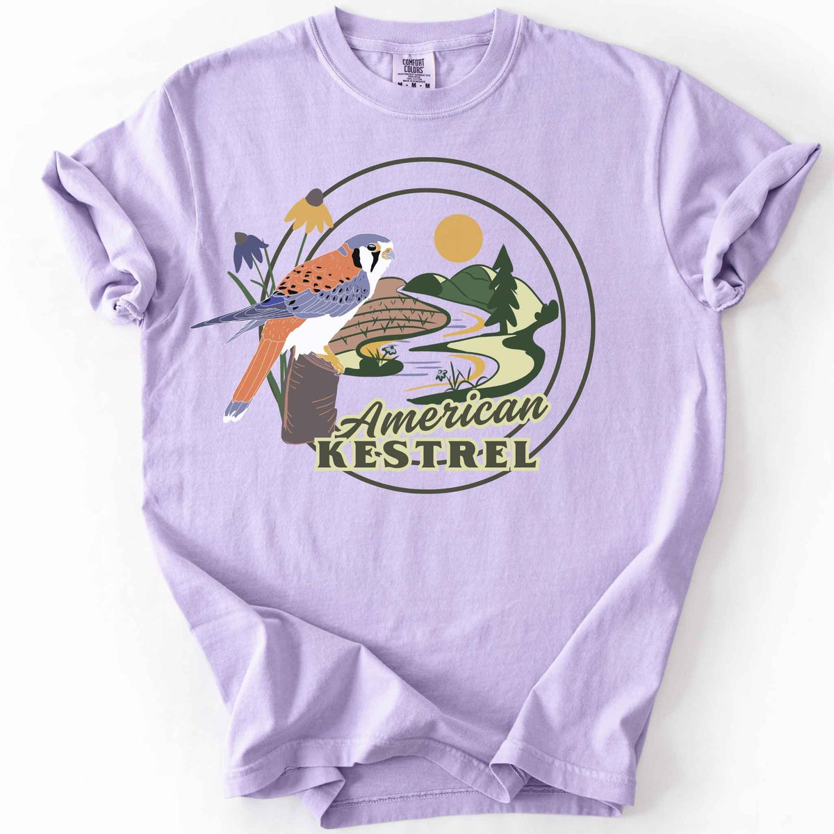 Orchid "American Kestrel" t-shirt on Comfort Colors tee, featuring a colorful illustrated kestrel perched among wildflowers with a farmland and river landscape in a circular badge design. Birdwatcher and raptor lover gift shirt for wildlife and nature enthusiasts.