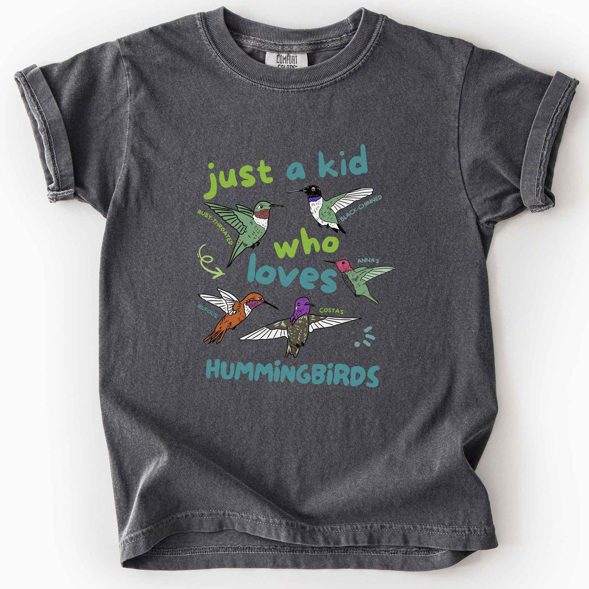 Pepper "Just a Kid Who Loves Hummingbirds" youth t-shirt on Comfort Colors tee, featuring five labeled hummingbird species including Ruby Throated, Anna's, and Costa's with playful green and blue lettering. Kids birdwatcher gift shirt for young nature lovers and budding wildlife enthusiasts.