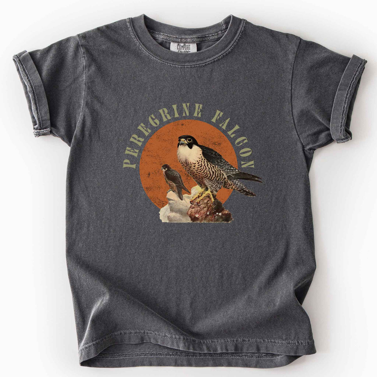 Pepper "Peregrine Falcon" kids t-shirt on Comfort Colors tee, featuring vintage style falcon illustration perched on a rock against an orange sun backdrop, with arched cream text. Birdwatcher and raptor lover gift shirt for falconry and wildlife fans.