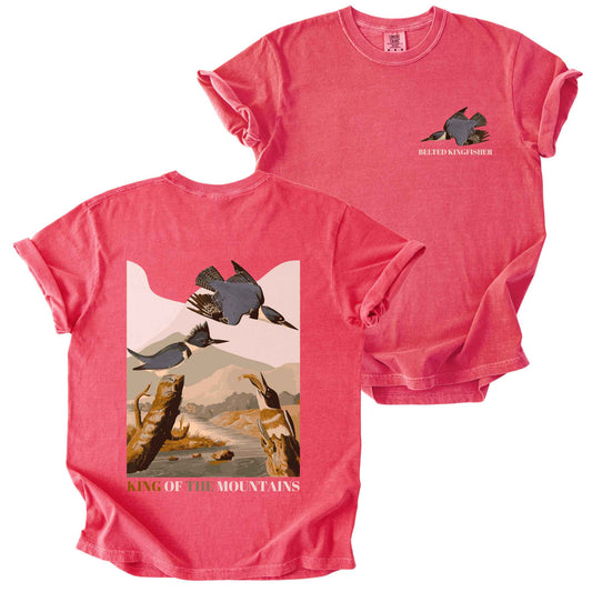 Watermelon "Belted Kingfisher, King of the Mountains" t-shirt on Comfort Colors tee, featuring a small front chest logo and a full illustrated back print of kingfishers over a river landscape in retro poster style. Birdwatcher gift shirt for wildlife and nature enthusiasts.