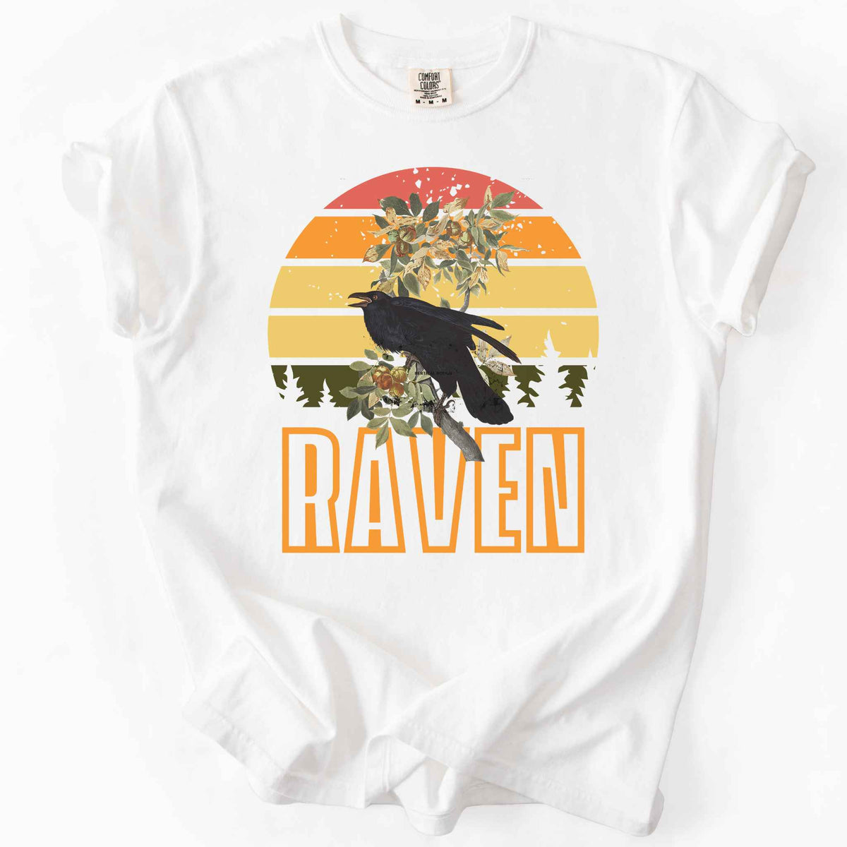 White "Raven" t-shirt on Comfort Colors tee, featuring a raven perched on a branch against a retro sunset sunburst design with bold orange text. Bird lover and wildlife gift shirt for corvid and nature enthusiasts.